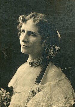 A vintage black and white portrait photograph of a woman with parted curled hair, wearing a white gown and a necklace of pearls or gemstones,