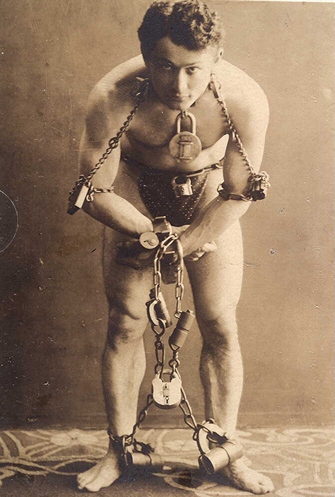 A sepia portrait photograph of a young man. He is standing, wearing only swimming trunks and is bound at the wrists, ankles, elbows and neck with chains and padlocks. The restraints mean he is standing in a hunched posture. He is looking directly at the camera.