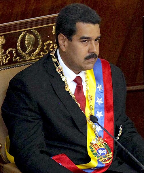 A photograph of Nicolás Maduro seated during an official ceremony.
A Hispanic man with short black hair and a moustache, he is wearing a suit, as well a decorated chain around his neck and a broad Venezuelan flag sash.