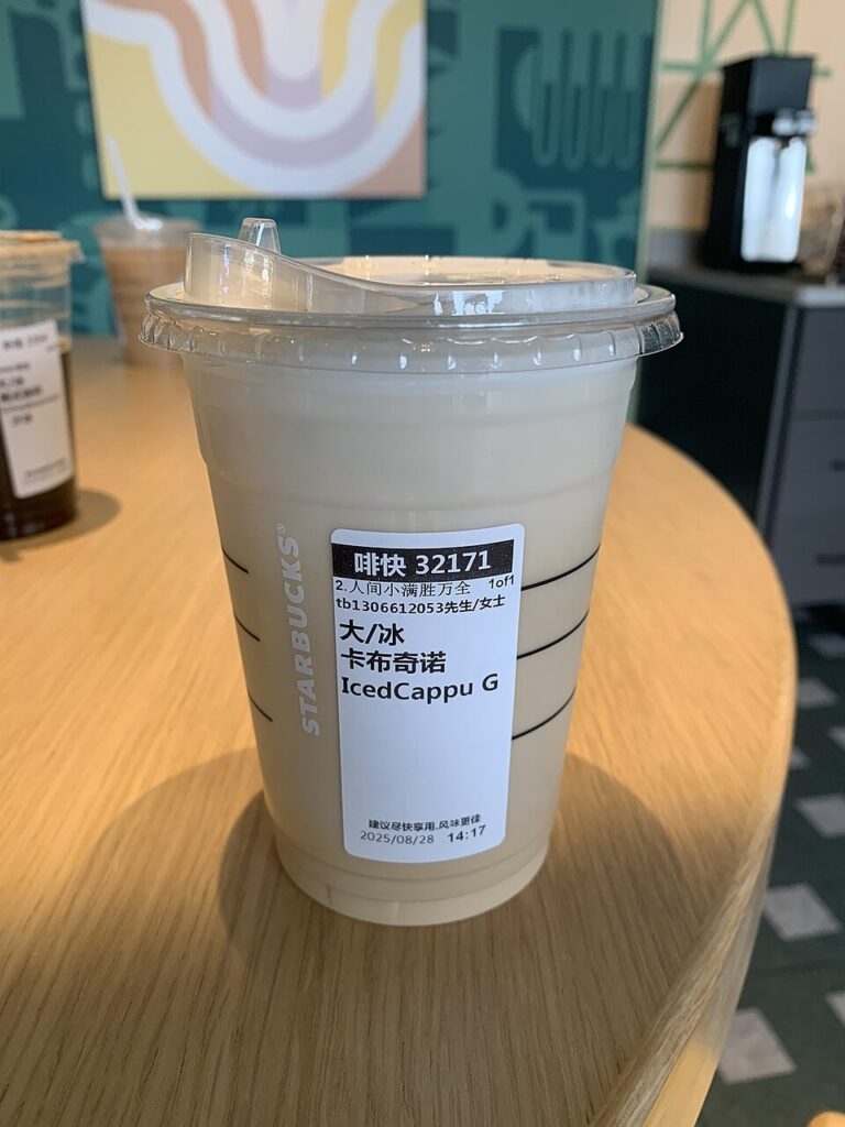 A clear plastic cup filled with a pale brown milky coffee drink. The cup has a clear plastic lid with a raised drinking spout for sipping from, somewhat resembling a child's sippy cup. The cup has STARBUCKS printed on it, and carries a label with some Chinese writing printed on it as well as "Iced Cappu".