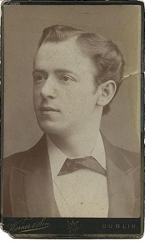 A vintage black and white portrait photograph of a clean-shaven man with parted short dark hair, wearing a white white shirt, dark neckcloth, and dark jacket.
