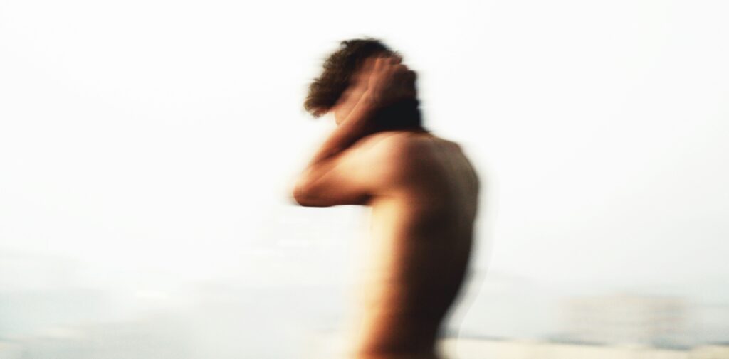 A deliberately blurred photo of a topless person, facing left, with their hands up alongside their head in an anxious sort of post.