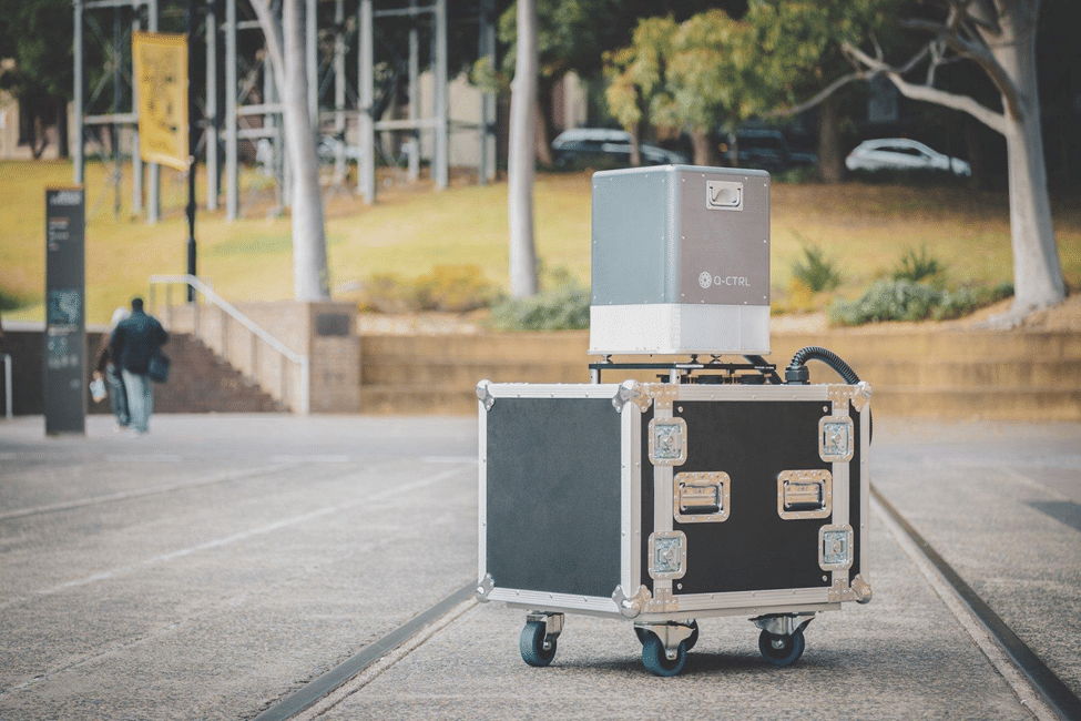 An image of an almost-cube black and silver kit box on wheels with another box atop it, mostly silver with some white, that has a Q-CTRL logo on its side and small metal handles. A plastic hose emerges from the lower box and we cannot see where it re-enters. It appears to be positioned in a car park on a college campus or similar location.
[Fair use: described in the text as analysis]