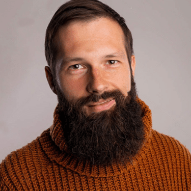 Profile image of a white, brown-eyed, brown-haired man with a large beard and short haircut, wearing a roll-neck thick-knit jumper in rust brown/orange. A featureless beige-grey background is behind. 