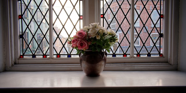 A window with diamond-shaped leaded panes. A vase of flowers sits on the windowsill and a street can be seen outside.