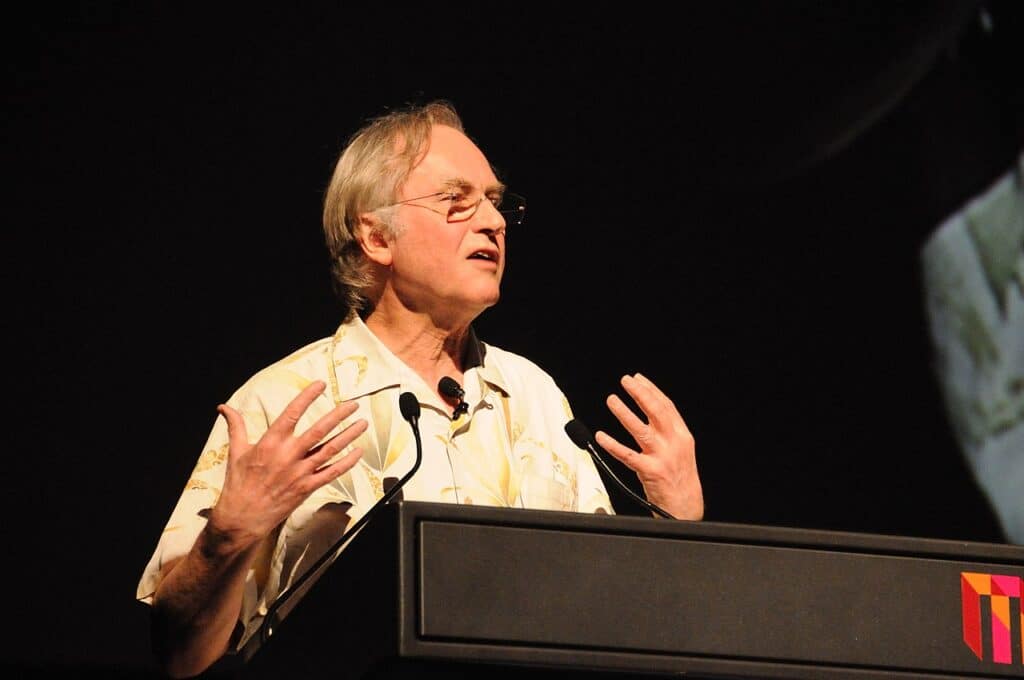 Richard Dawkins stands at a podium with microhones, speaking and gesturing with his hands against a black background