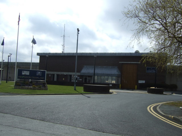 A photograph of the driveway to an austere low-rise building with lights and cameras on poles, and a sign saying "HM Prison Frankland".