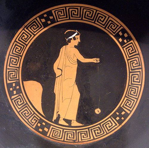 A black square piece of ancient ceramic bearing an orange-red image of a boy wearing a toga, standing with his right hand held in front of him and a disc hanging from his hand by a string.
The boy is framed by an orange-red circular design containing dots and spirals.