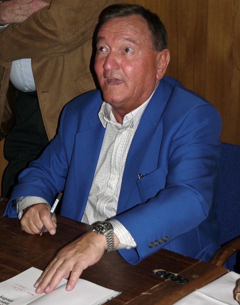 Author Erich von Däniken sits at a desk, wearing a sapphire-blue jacket over a white shirt, and a wristwatch, having just signed a book