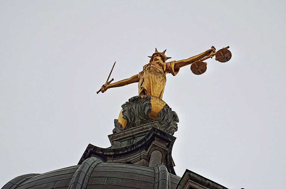 A photograph looking up at a gilded statue of 'Lady Justice' atop a courthouse. She stands robed, arms held straight out to her sides. Her right hand holds a sword pointing up, her left hand holds a set of weighing scales.