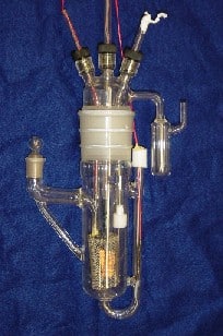 A complex piece of laboratory glassware, with metal leads running into it and some metal components inside. The item has a main hollow cylinder, with three plugged apertures at the top through which metal wires or pipes pass. What looks like a piece of bright silvery metal mesh sits inside the cylinder, connected to at least one of these three external probes. Three narrow glass tubes branch from the main cylinder. The size of the whole assembly is unclear, but perhaps on the order of tens of centimetres.
