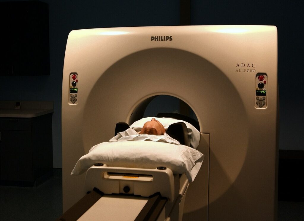 A person lies on a bed which is enclosed by a large douhgnut-shaped piece of machinery, with a few buttons on the otherwise blank face of the machine. Only the person's head is visible.