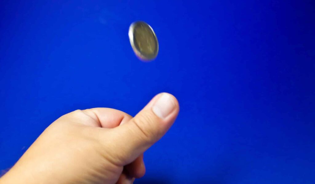 Against a sea-blue background, a white person's hand flips a silver coin with their thumb, it's mid-air in the image.