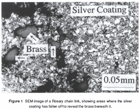 A black and white annotated photograph, with the caption "Figure 1. SEM image of a Rosary chain link, showing areas where the silver coating has fallen off to reveal the brass beneath it." The majority of the imaged area is pale and grainy, labelled "Silver Coating". Smaller smooth darker patches, labelled "Brass" are scattered about the surface. The imaged area measures approximately 0.25mm by 0.15mm according to a superimposed scale bar.