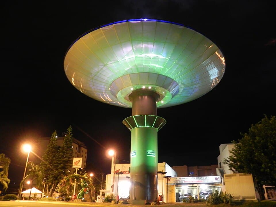 A photograph taken outside at night, looking up at an illuminated water tower, with a broad disc-shaped reservoir atop a single relatively narrow column.
