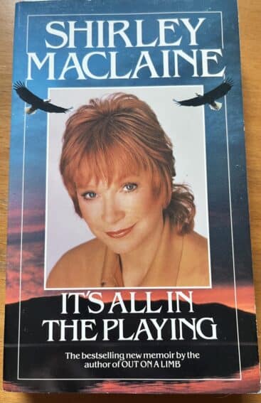 A photograph of the cover of a book.
In the centre of the cover is a portrait of a white woman with medium length red hair.
Above her is the author's name – "Shirley Maclaine".
Below her is the book's title – "It's all in the playing" and subtitle "The bestselling new memoir by the author of OUT ON A LIMB"
A pair of soaring eagles are superimposed just under the author's name.