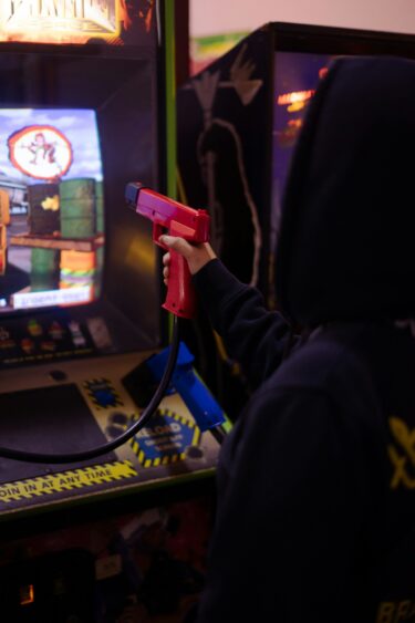 Someone playing an arcade video game. The screen shows a scene with oil barrels and what might be explosions or gunfire. The player is holding a controller shaped like a pistol, connected to the game unit by a cable.