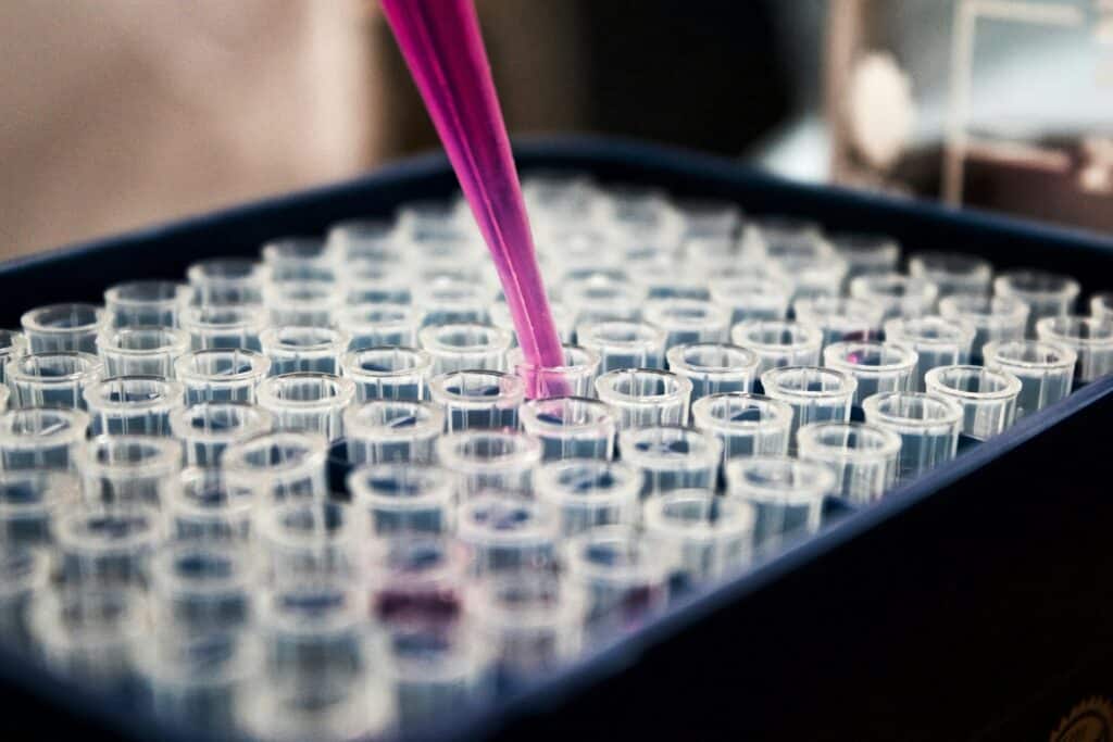 A photograph of a tray containing an eight by twelve array of 96 small glass tubes. A purple liquid is being added into one of the tubes by a pipette.