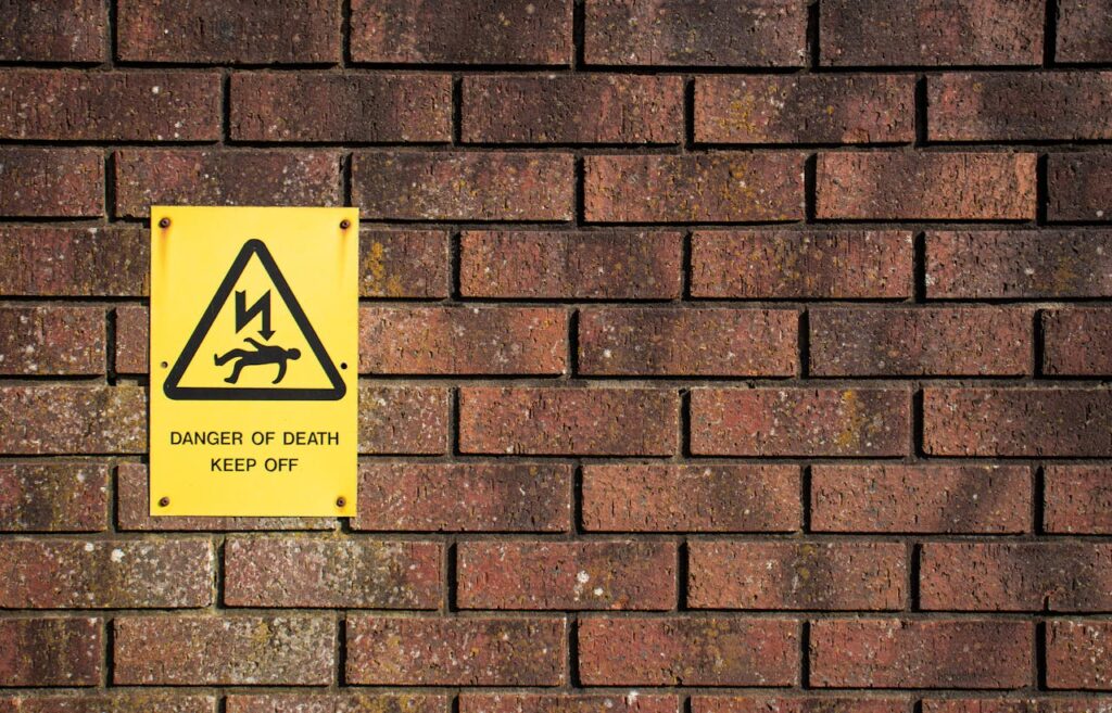 A photograph of a brick wall bearing a yellow hazard warning sign.
The sign reads "Danger of death. Keep off", under a warning triangle in which a stick figure on the lying ground appears to have been struck by a bolt of electricity.