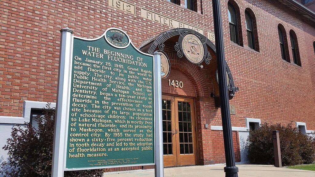 A photograph of part of the outside of a large brick building with FILTER PLANT in large embossed letters over the door. A historical information sign is nearer the camera.
The sign reads
"The Beginning of Water Fluoridation
On January 25, 1945, Grand Rapids became the first city in the world to add fluoride to its public water supply. The city, along with the US Public Health Service, the Michigan Department of Health, and the University of Michigan School of Dentistry, began a ten year study to determine the effectiveness of fluoride in the prevention of tooth decay. The city was chosen as a test site because of its large population of school-age children, its closeness to Lake Michigan which is mostly free of natural fluoride, and its proximity to Muskegon which served as the control city. By 1955 the study had shown a sixty-five percent reduction in tooth decay and led to the adoption of fluoridation as an accepted public health measure."