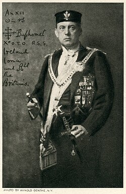 A black and white portrait photograph of a standing man, showing him from the knees up. He is looking directly at the camera and dressed in a white shirt with formal dark frock coat, and on his head is wearing a dark cylindrical cap. The cap has a winged badge on it, and there are many medals and decorations on the shirt and coat. A wide ornamental chain or ribbon hangs around the man's neck. In his right hand he is holding a short thin staff or cane, and a sword hangs at his left hip by his left hand. Some handwritten text and symbols on the left hand of the photograph are as follows: An XII ☉ in ♑ [cross] Baphomet X° O.T.O. R.S.S. Ireland Iona and All the Britains.