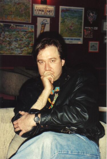 Comedian Bill Hicks at the Laff Stop in Austin, Texas. He wears a leather jacket and jeans, and holds his fingers over his mouth thoughtfully, while his left arm is across his body.