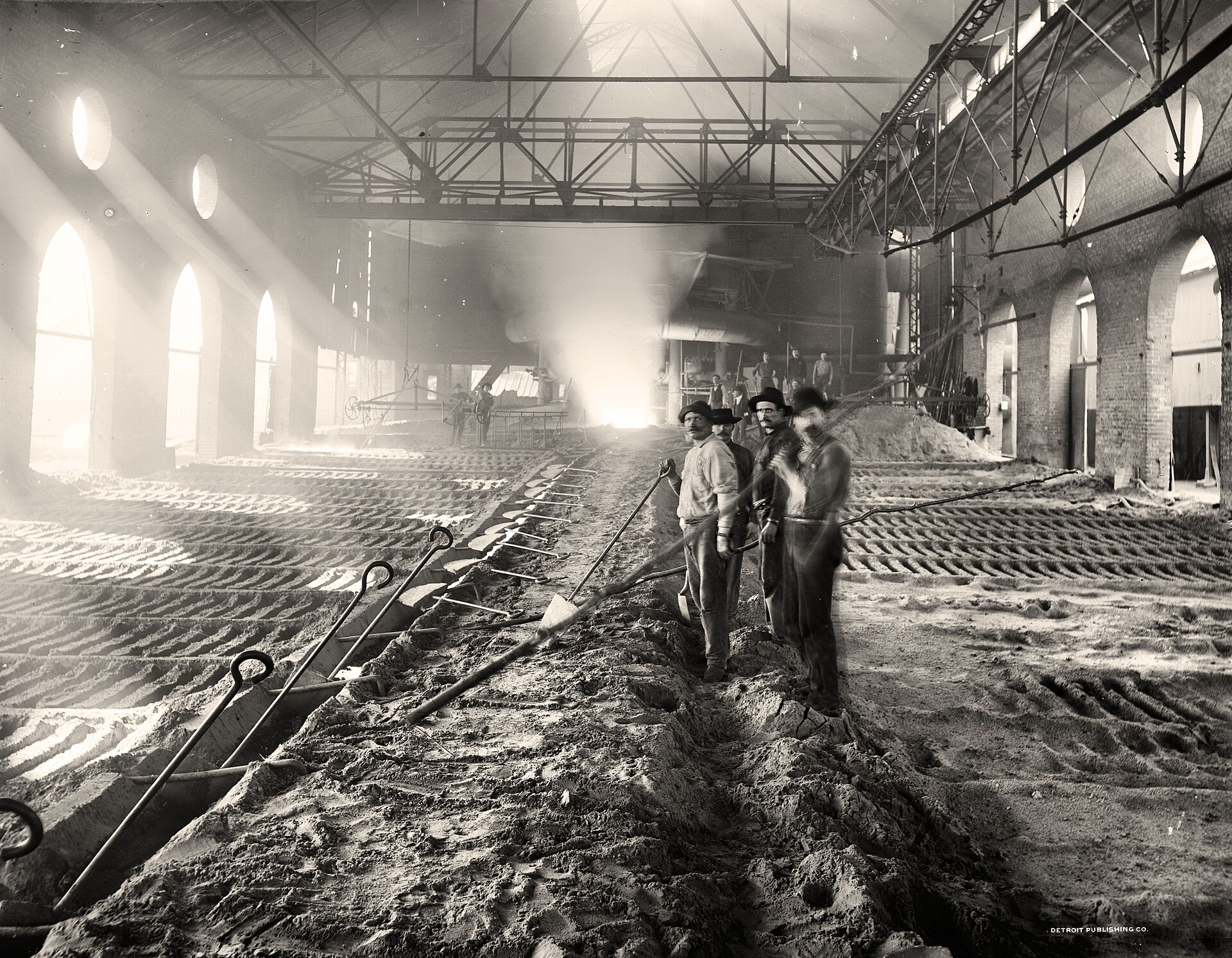 Four iron-worker men stand in the centre of a large factory space with light streaming in from windows on the left and girders supporting the ceiling above. They hold long implements and stand in sandy material. Other men are visible in the background of the same factory hall.