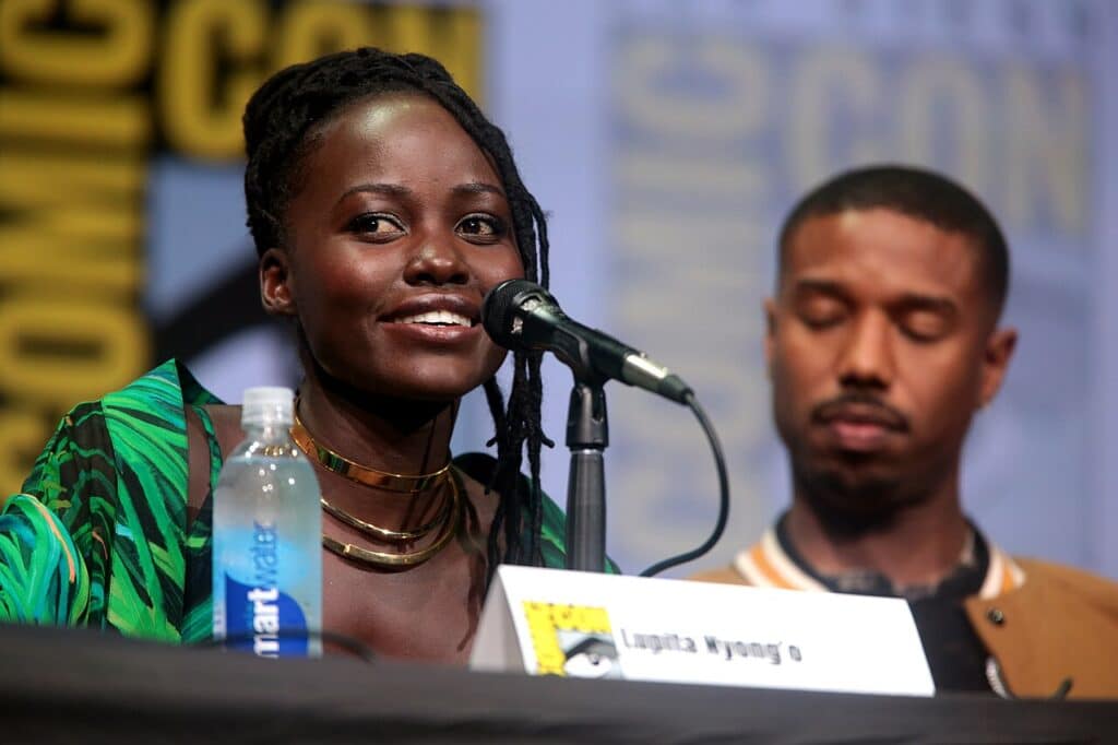 A photograph of a young Black woman sat at a table with a microphone in front of her, and a name plaque saying "Lupita Nyong'o". A Black man is slightly out of focus seated further from the camera.
In the background is banner reading "Comic Con"