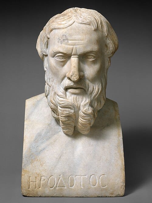 A photograph of a marble bust of a man with a stern expression and curly beard. Greek text carved into it reads "Hrodotos".
