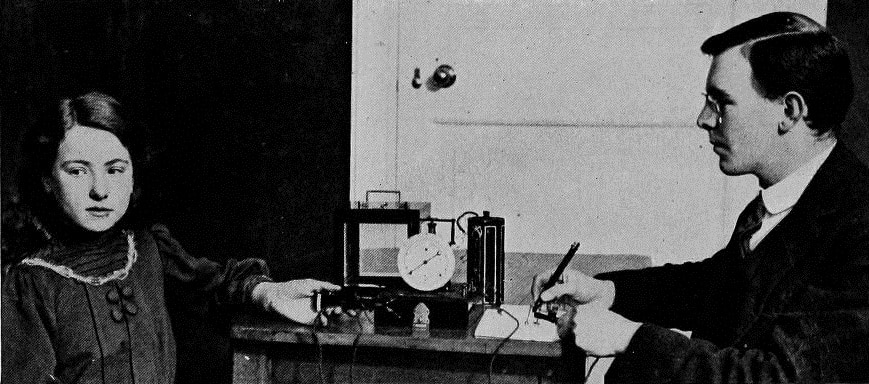 A grainy black and white photograph showing two people sitting opposite each other at a desk. On the left hand side is a girl of maybe 10 years. On the right hand side is a young man. The man is looking at the girl, and writing in a notebook. The girl is looking away from the man, and one of her hands is resting on the desk, touching an apparatus composed of boxes, wires, and a prominent dial.
