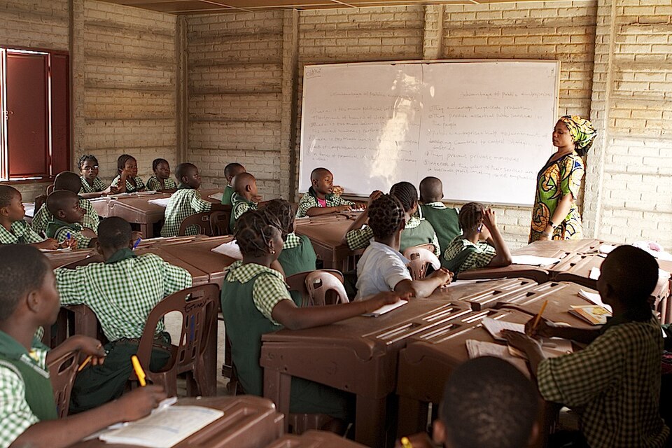 Photograph taken inside a schoolroom. A Black woman wearing a boldly printed dress and matching headwrap stands at a whiteboard.
Approximately twenty Black boys and girls, wearing green and white chequered uniform shirts, sit at desks and are looking toward the teacher. 
