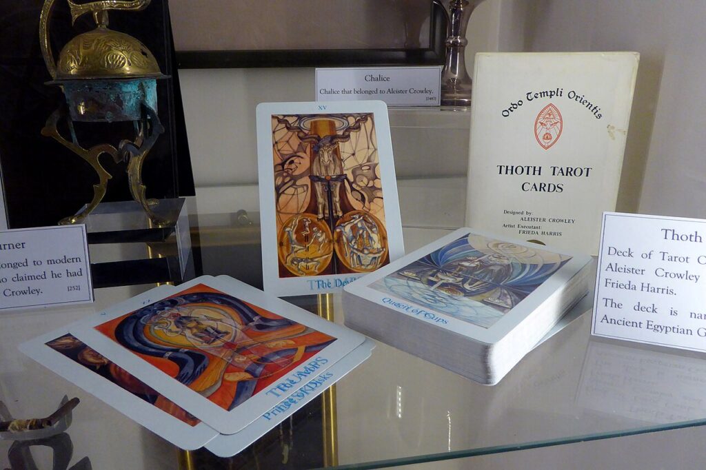 A photograph of a deck of cards on a glass shelf, arranged so the faces of three cards are visible. The visible cards are "The Devil", with an illustation of a long-horned goat, "The Aeon", with an illustration of someone seated among swirls of colour, and "Queen of Cups" with an illustration of a seated woman pouring water from a cup. Next to the cards is the box for the deck, bearing the words "Ordo Templi Orientis" and "Thoth Tarot Cards". Under this text is "Designed by: Aleister Crowley. Artist executant: Freida Harris" In the background is a different shelf with a silver cup next to a card reading "Chalice that belonged to Aleister Crowley".