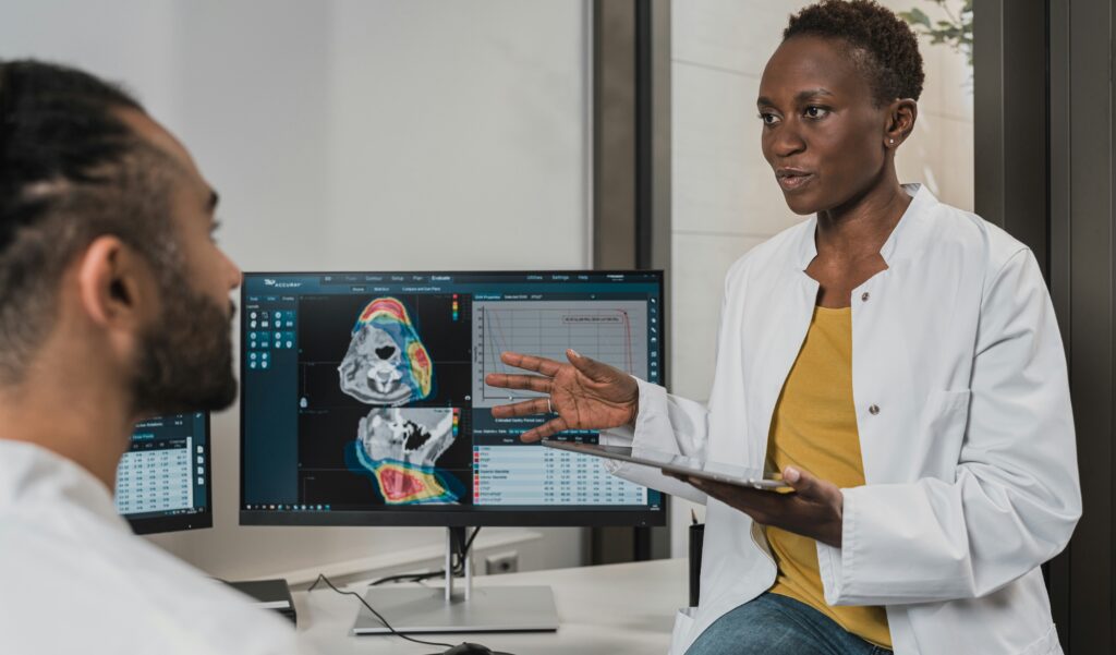 A doctor in a white coat over a mustard top and jeans half-sits on a table and holds a clipboard while explaining something to a colleague or patient in front of them. The doctor is a Black woman and opposite her is a man. The computer screen shows some medical imagery