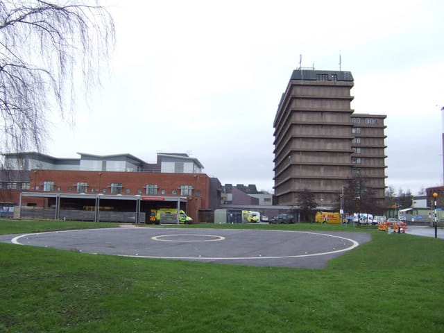 A photograph of a set of buildings under an overcast sky. To the left is a three storey brick building, with an ambulance under a portico. To the right is a nine or ten storey concrete tower with windows. A round concrete helipad is set into the grass between the buildings and the camera.