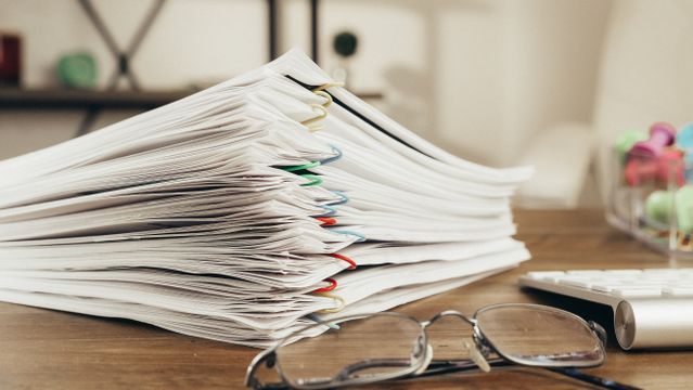 A photograph of several sets of papers, paperclipped together and neatly placed on a desk, next to a pair of spectacles and a keyboard.