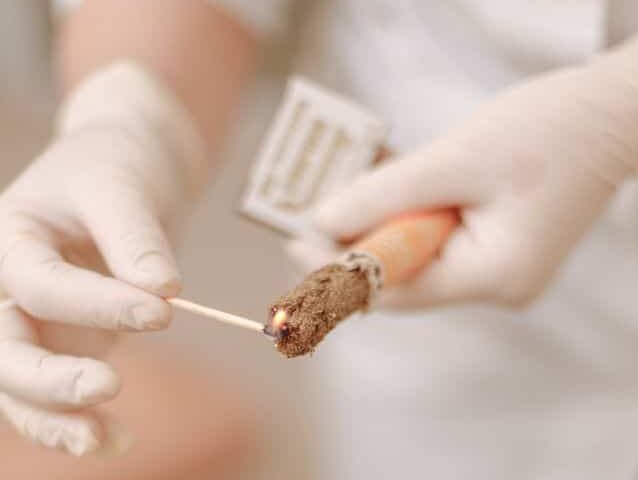 A photograph of someone's gloved hands as they touch a burning a match held in their right hand to a cigarlike roll of dried leaves held in their left hand.