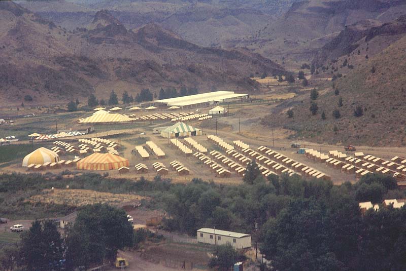 A photograph taken outside from an elevated position looking onto a flat expanse of ground set among steep hills. The flat area contains maybe a hundred huts and several large marquee-style tents.