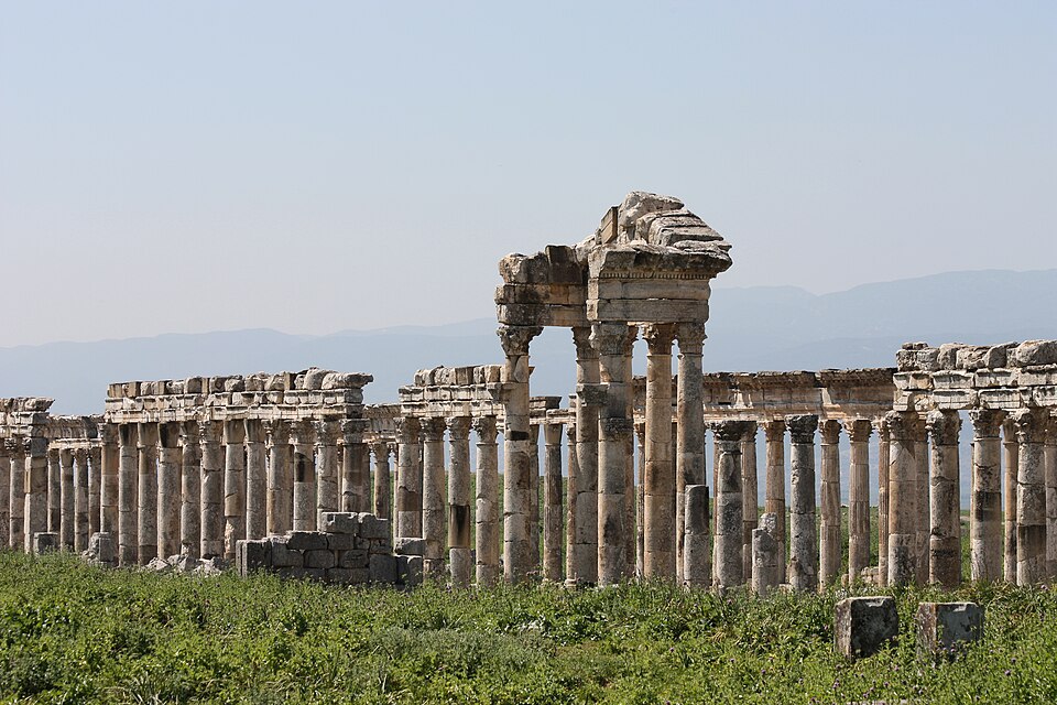 A colour photograph of a two or three long parallel lines of old stone columns, many supporting weathered remains of roof masonry. They rise from flat scrub-covered ground, and hills rise in the distance.