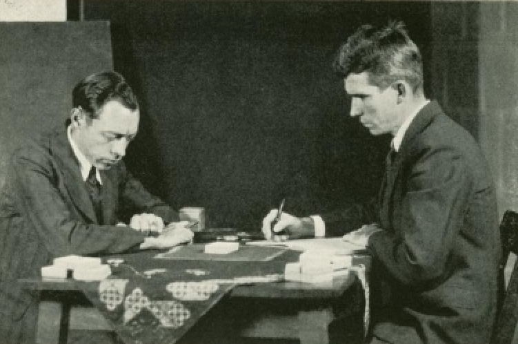 Black and white photograph of two white men wearing suits, seated across a table from one another. The man at left appears to be concentrating on a small pile of cards in the table. The man at right is making notes in a book.