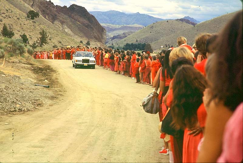 A photograph taken outside, showing a white Rolls Royce car driving along a dirt road. Along one side of the dirt road stand dozens of people wearing red, dark orange, and purple clothes.
The landscape beyond is steep hills dotted with a few trees.