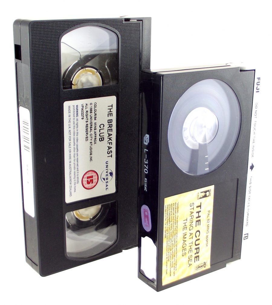 A colour photograph of two plastic-bodied tape cassettes side by side. At left is a VHS cassette, which is approximately 25% longer than the Betamax cassette beside it.
