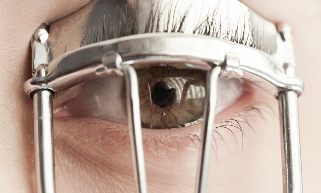 A close-up of a white person's hazel-irised eye, they're using eyelash curlers and the visual effect created by the curlers' vertical metal bars is of a prison cell