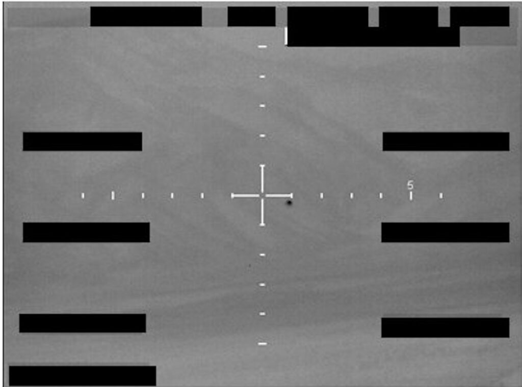Still black and white image from a video feed, looking down onto a landscape of light and dark fields. A white crosshair is marked in the middle of the image and there are many black rectangles, presumably redacting onscreen data. Just below and to the right of the crosshair is a tiny black circular object lacking any detail.