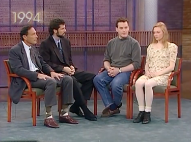 Four people sitting in chairs on a television set. From left to right: A clean-shaven man with black hair wearing jacket and tie. a bearded man with black hair wearing jacket and tie, a casually dressed clean-shaven young man with brown hair, and a casually dressed blonde-haired young woman.