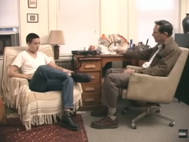 A still frame from a video, depicting an interior scene. A young woman with short black hair wearing t-shirt and jeans is seated in an armchair. At a nearby desk a middle-aged man with black hair is seated in a swivel chair, turned to face the woman as if in conversation.