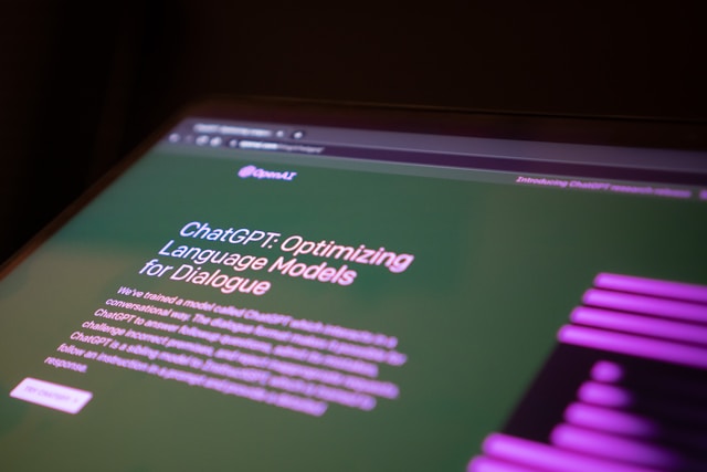 A photograph of a laptop screen OpenAI logo and the title "ChatGPT: Optimizing Language Models for Dialogue." Under this is some partially blurred text readingW"e've trained a model ChatGPT which [blurred] conversational way..."