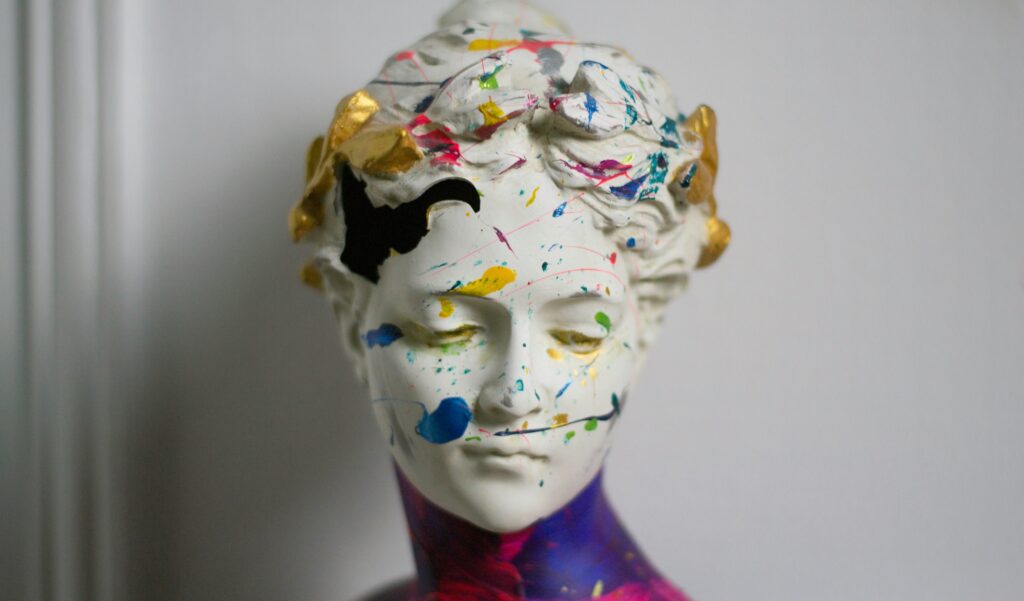 The head of a Greek-style statue of a woman, she is turned to her left slightly and looking downwards. There is a broken area around her forehead, showing the statue is hollow. Paint in various colours is splashed across her face and hair, her neck and shoulders in blue-purple and pink