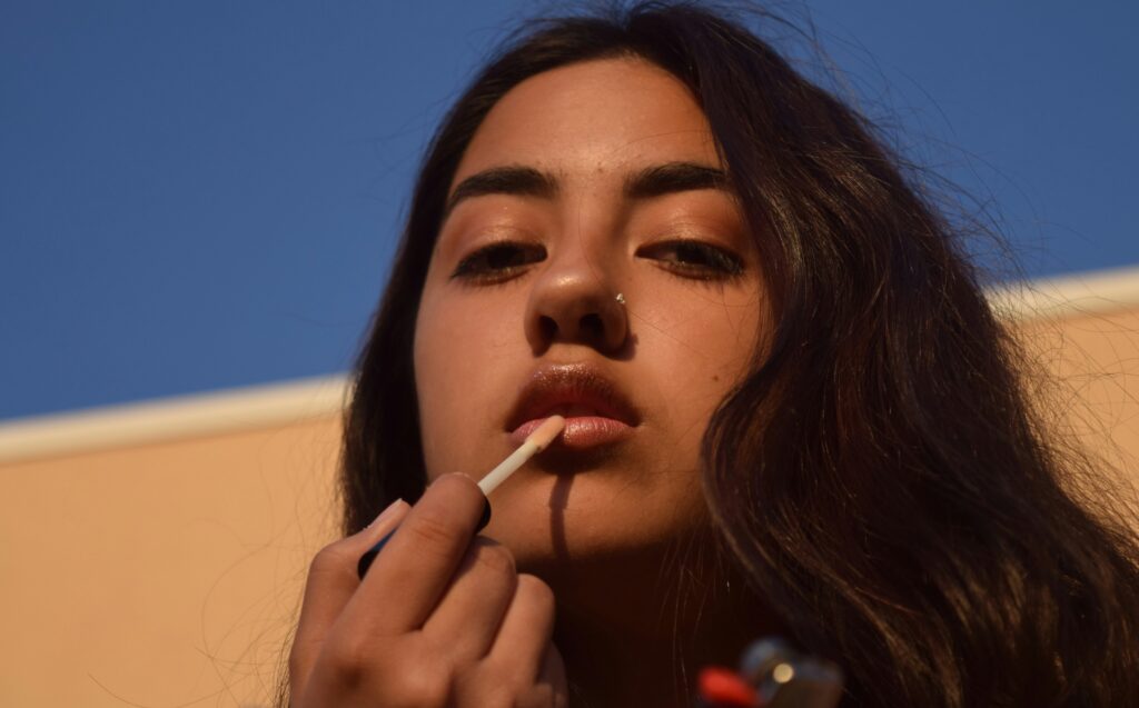 Close up on the face and hand of a Mid or West Asian woman applying nude lipgloss, standing outside with blue sky above her and the slanted lines of a building cutting the background. Her dark hair is loose and she looks down at the camera
