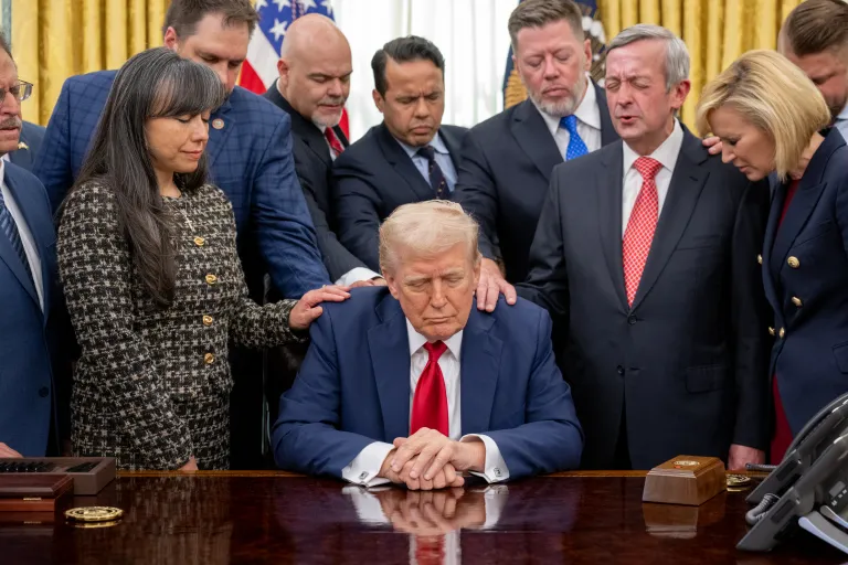 Photograph of Donald Trump sat at the Resolute Desk, eyes closed. Standing beside and behind him are several men and women, with their eyes closed, hands extended to rest on Trump's shoulders.