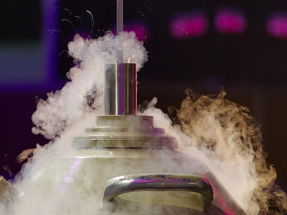 Photograph of the upper part of a cylindrical metal storage canister, with a small opening at the top. It is wreathed in a cloud of mist, formed as water vapour in the air freezes.